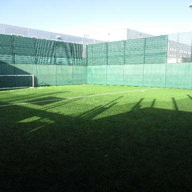 Photograph of a small outdoor football field enclosed by green fencing and surrounded by buildings. The field features artificial turf, a single goalpost, and visible white boundary lines with shadows cast across the playing surface.