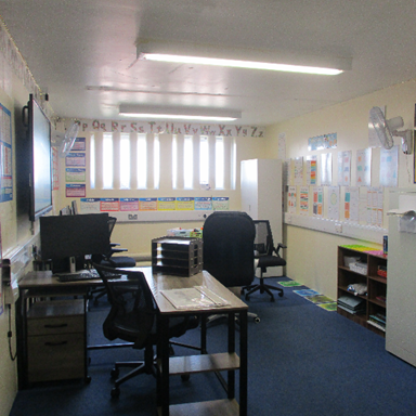 Photograph of a small classroom with desks, chairs, and computer monitors. Walls decorated with educational posters, alphabet letters, and storage units.