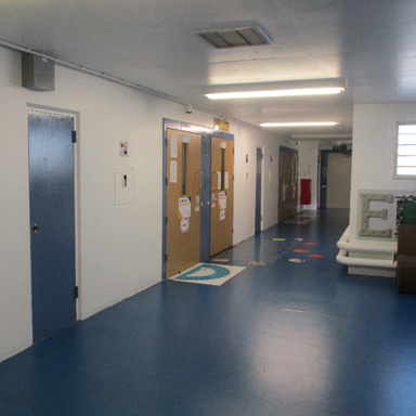 Photograph of an indoor hallway with blue flooring, white walls, and multiple closed doors, some with notices attached.
