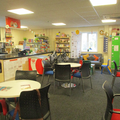 Photograph of a visits area featuring multiple round tables with black and red chairs arranged around them. The room includes a counter with snacks and beverages, shelves stocked with various items, colourful cushions on a window seat, and bright overhead lighting.