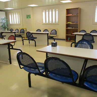 Photograph of a waiting room with multiple rows of blue plastic chairs attached to black metal frames and white rectangular tables. The room has beige walls, windows with vertical blinds, a wooden bookshelf with books, and numbered table markers.