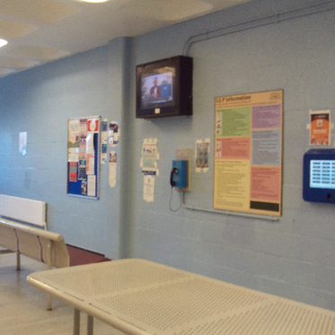 Photograph of the holding area, which is clean and bight. There is a TV and information posters on the wall, and some fixed metal benches and a table.