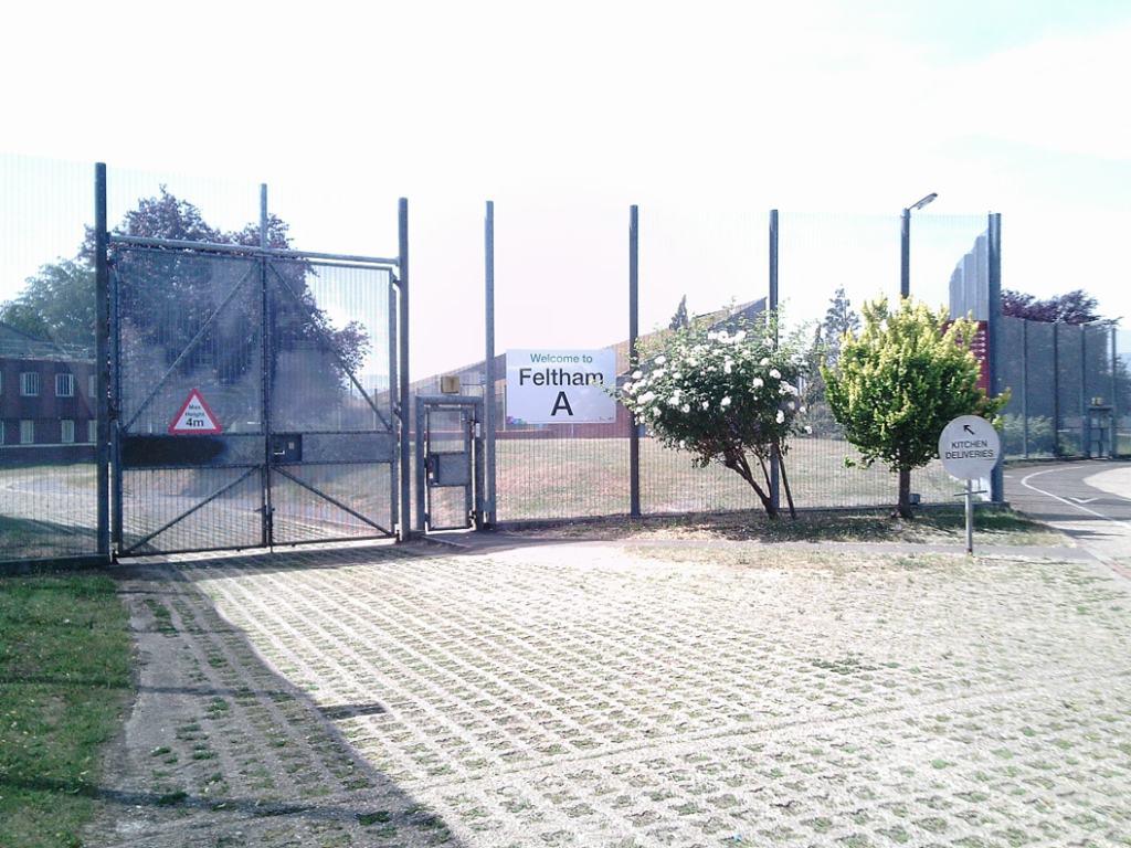 Photograph of the outside of Feltham A, showing a collection of low buildings behind a high perimeter fence.
