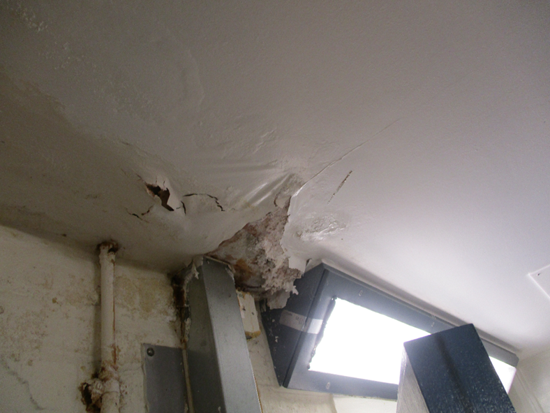 Photograph showing a damaged ceiling corner with peeling paint, cracks, and exposed underlying material near a metal beam and a bright light fixture.