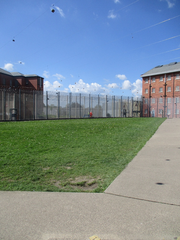 Photograph of an outdoor prison yard enclosed by tall fences topped with barbed wire, with brick prison buildings on either side under a clear blue sky. The yard features a grassy area and a concrete walkway, highlighting security measures and institutional environment.