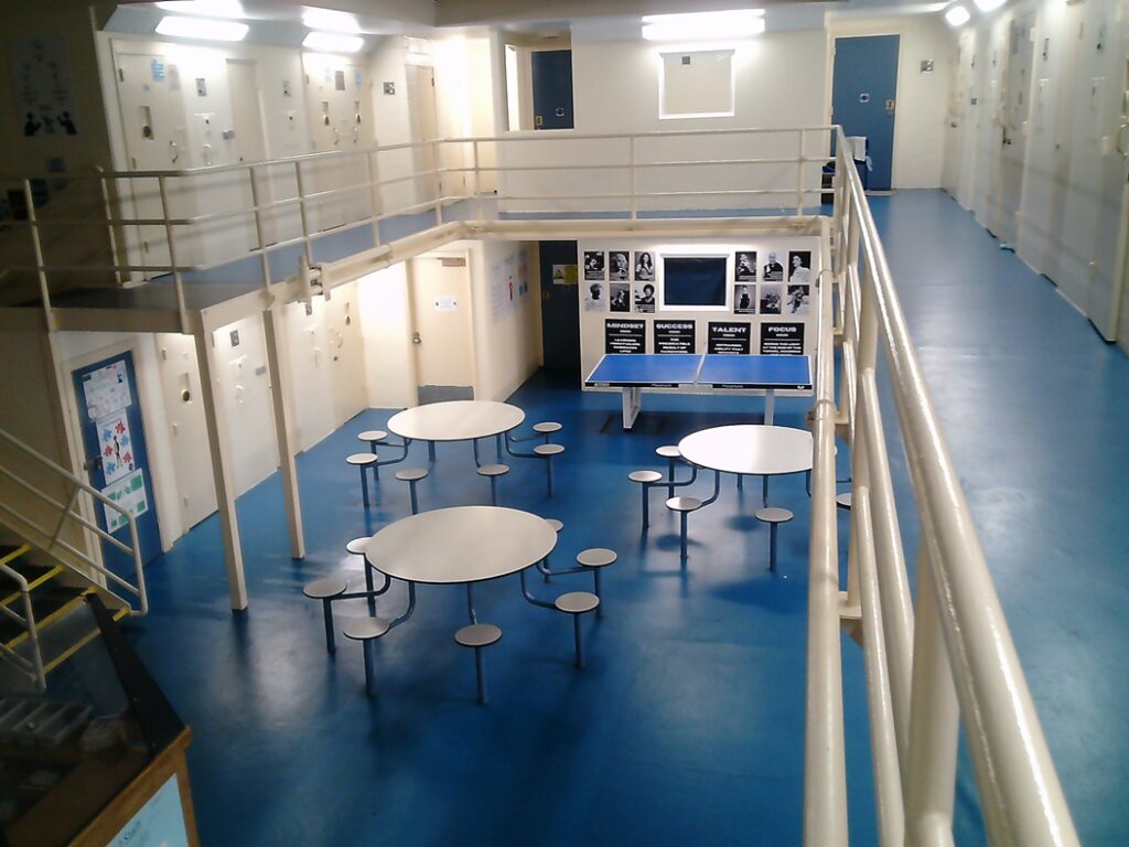 Photograph of an indoor recreational area featuring blue flooring, round tables with attached stools, and a ping pong table against a wall with black-and-white posters. The space is surrounded by white cell doors on two levels connected by metal railings and stairs.