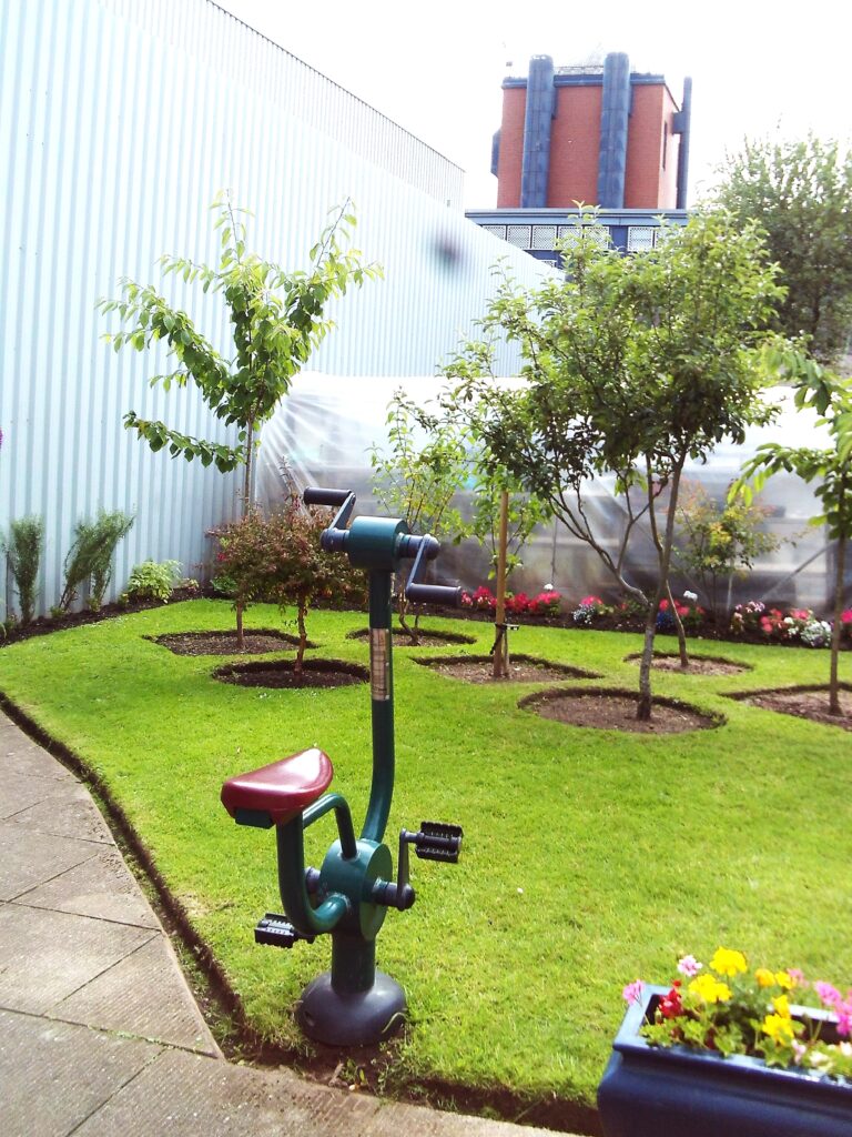 Photograph of the J wing garden with several trees and some flower beds, and an exercise bike visible.