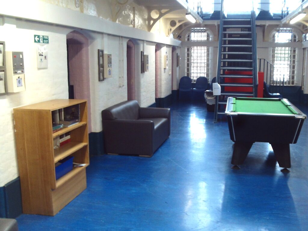 Photograph of a communal area on D wing. A bookshelf, pool table and sofas can be seen, and stairs lead to an upper floor.
