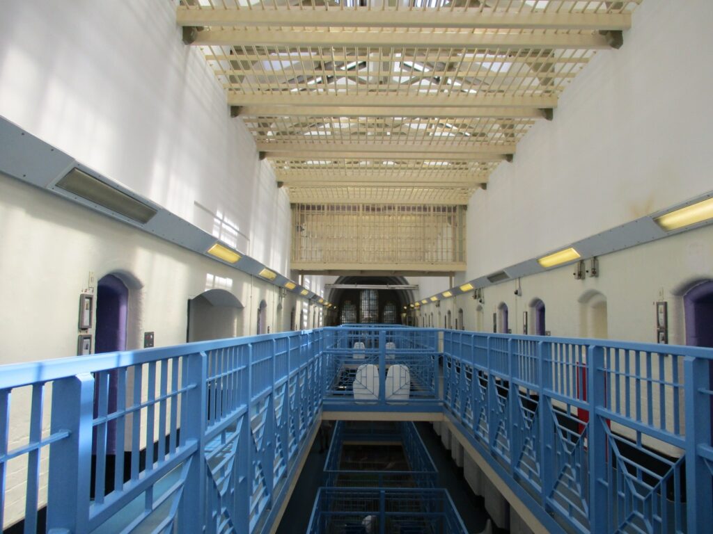 photograph of a wing landing inside HMP Birmingham. The landing has blue banisters and doors can be seen off both sides of the landing.