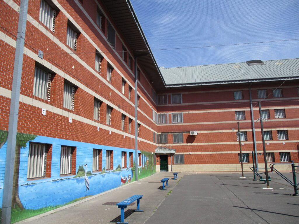 Photograph of an exercise yard enclosed by prison buildings. The brickwork on the ground floor has been decorated by a mural and some seats and exercise equipment arranged in rows in the yard.