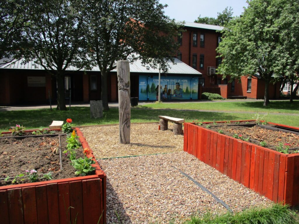 Photograph of some raised beds that have been added to the visitor centre garden. There is evidence of planting in the beds, and a bench, trees and buildings can be seen in the bacxkground.
