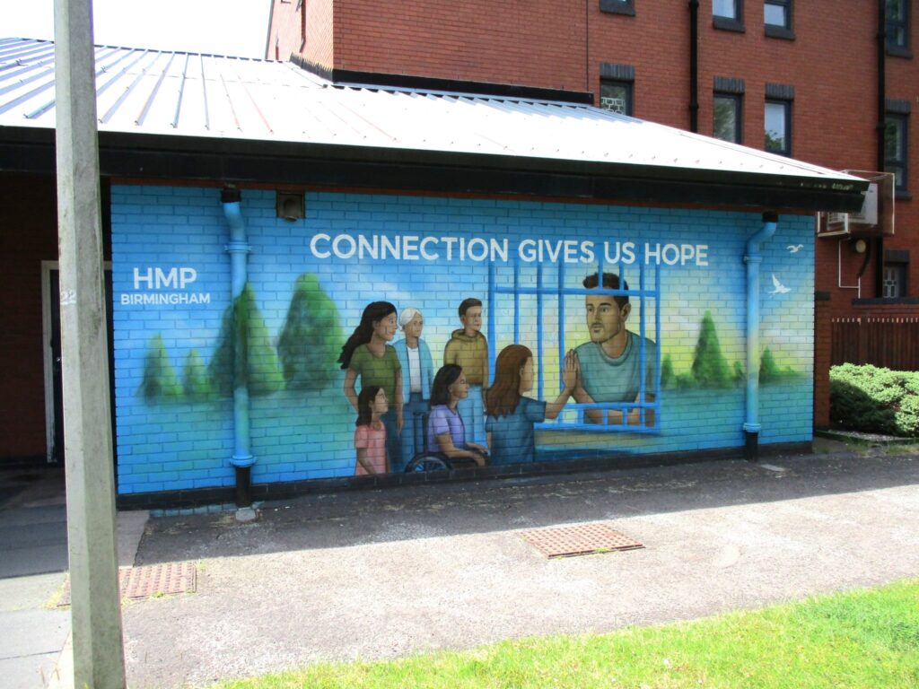 Photograph of a mural that has been painted on the side of a building in the visitor centre grounds. It features a prisoner behind bars, with family and friends looking at him, and at the top of the image there is writing that says "Connection gives us hope".