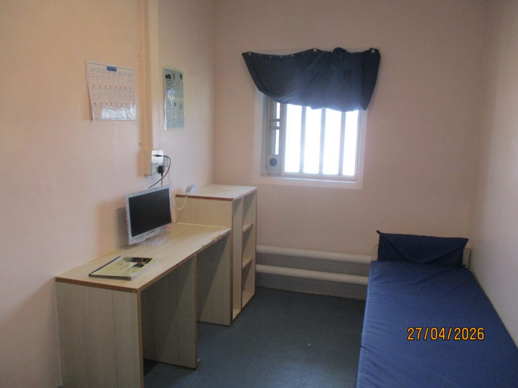 Photograph of a first night cell ready for occupation. A bed is one side of the room and a desk and storage furniture are on the other side. There is a window in the back wall.