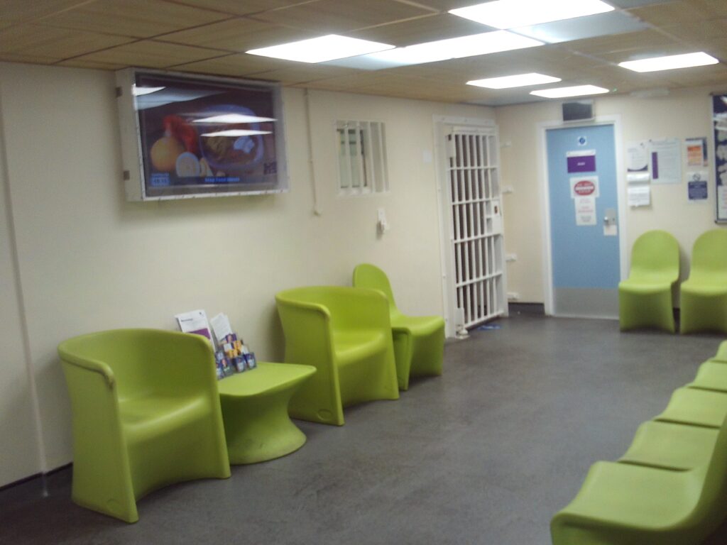 Photograph of the health care waiting area. Green moulded chairs are arranged around the perimeter of the room and a creen is mounted on one wall.