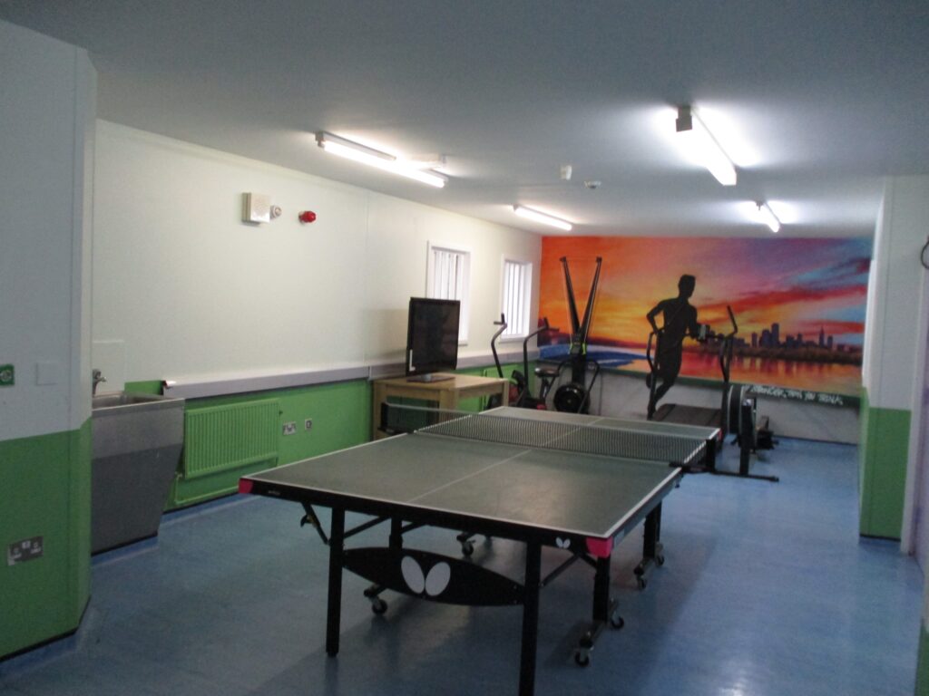 Photograph of the ISFL Association room. A table tennis table can be seen in the foreground. Behind it there is a table with a television on it and some exercise equipment.