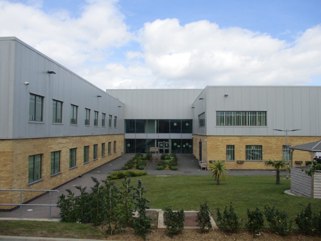 Photograph of the exterior of HMP Five Wells, showing a large section of a two-storey building with windows on both levels, and a grass area with some shrubs and bushes planted in the foreground.