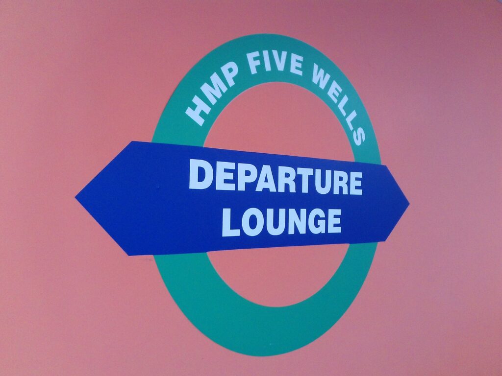 Photograph of the 'HMP Five Wells departure lounge' sign, a graphic based on the roundel design for the London Overground, but rendered in different colours.