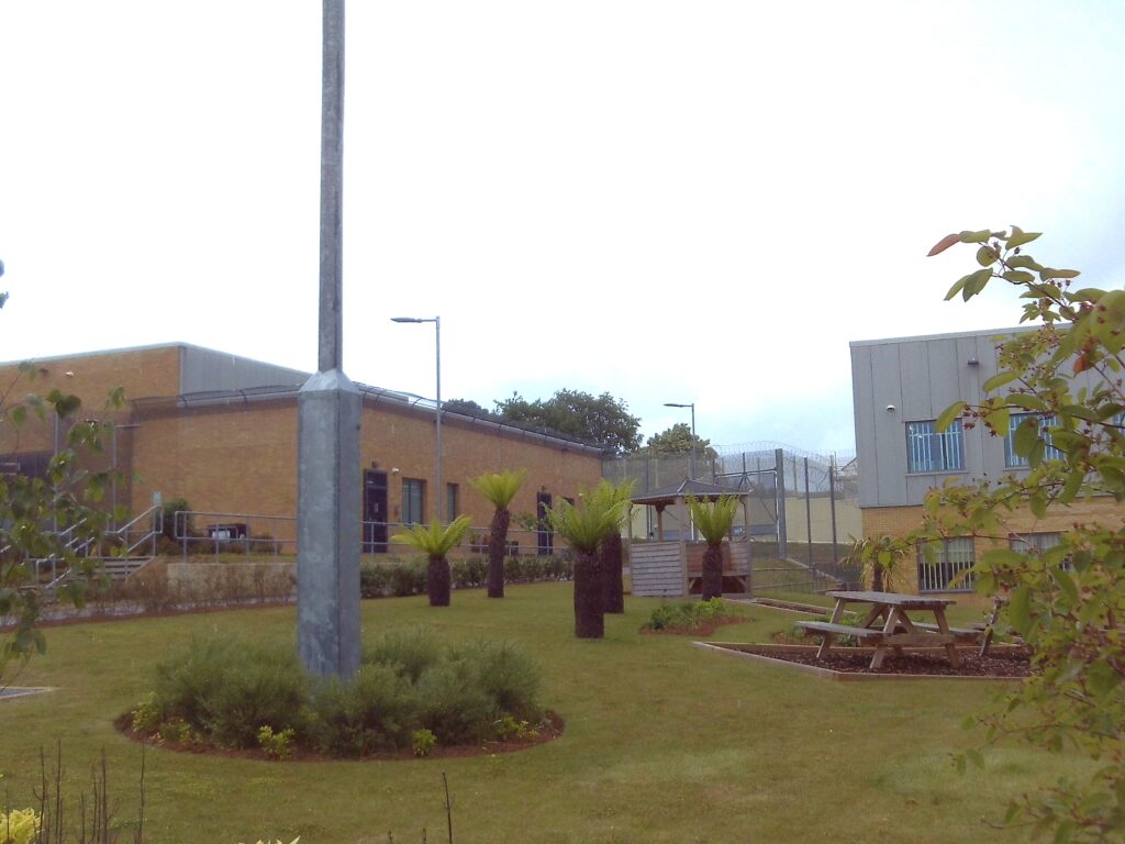 Photograph of outdoor space, with trees, shrubs, grass and a picnic table, with prison buildings in the background.
