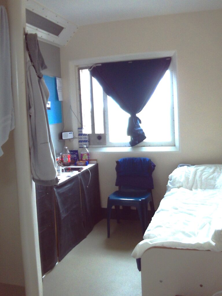 Photograph of a single cell, with a bed against one wall, a chair under the window, and storage furniture on the other side.
