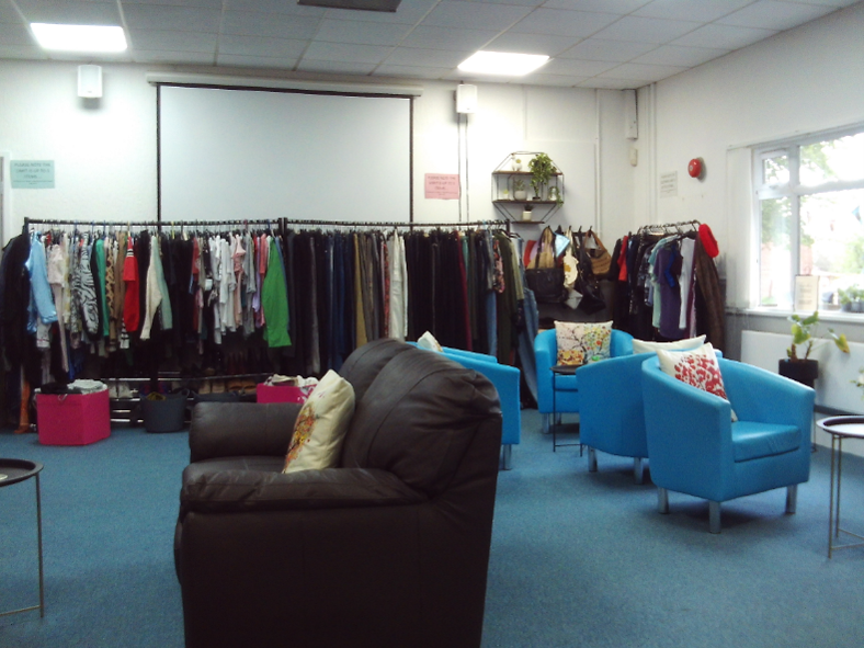 Photograph of the departure lounge, showing a room with some rails and boxes of clothing, a sofa and some armchairs facing small coffee tables, and some potted plants.