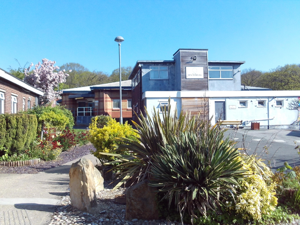 Photograph of the exterior of New Hall, showing shrubs and planting in the forefront, with a two-storey building behind. To the left is a path leading to the building, and to the right is a driveway.