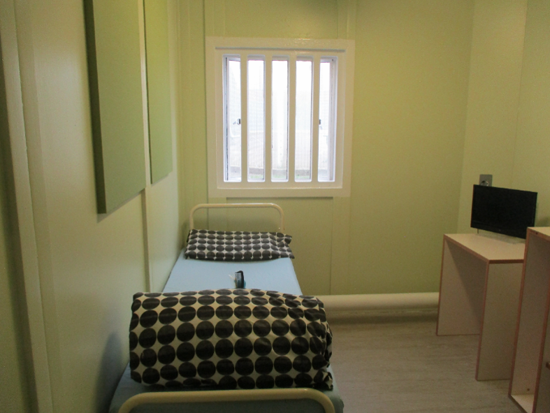 Photograph of a first night cell, showing a clean and tidy room, with a bed, a desk, a small TV and a storage unit.