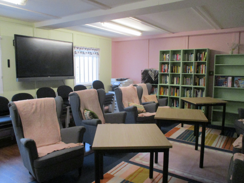 Photograph of the induction room. There are two rows of armchairs, with three coffee tables in between. There is a large TV on the wall, a stack of seats underneath it, and two large bookcases with books and games.