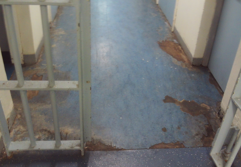 Photograph of a worn corridor floor with peeling blue linoleum and visible patches of exposed underlying material, with metal bars on the left and right sides.