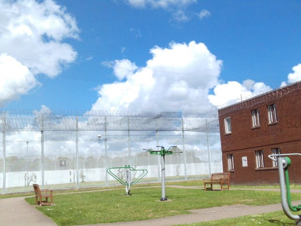 Photograph of an outdoor prison exercise yard featuring green exercise equipment, wooden benches, and a brick building on the right. The yard is enclosed by tall fences topped with barbed wire under a partly cloudy blue sky.