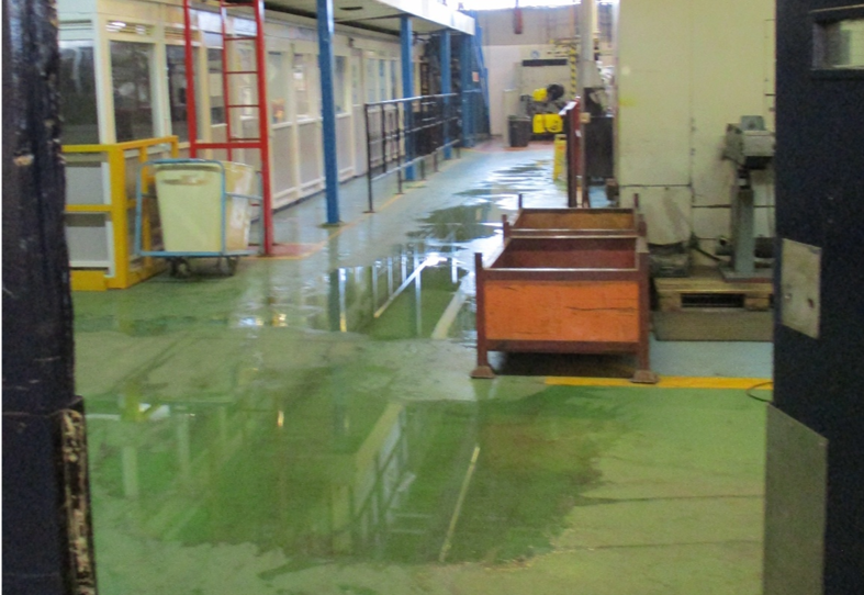 Photograph of an industrial workshop or factory floor with wet green concrete flooring and various equipment, including a red elevated platform and storage bins. Overhead fluorescent lighting and metal railings line the space.