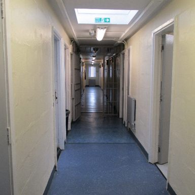 Photograph of a narrow corridor inside a prison, featuring metal cell doors on both sides and a blue floor. Overhead fluorescent lights and an emergency exit sign are visible, with a barred gate halfway down the hallway.