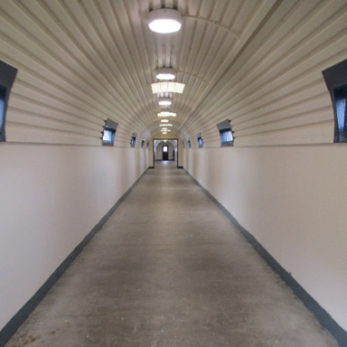 Photograph of a long, narrow corridor with a curved ceiling and evenly spaced ceiling lights. Walls are plain white with small windows on both sides, leading to a door at the far end.