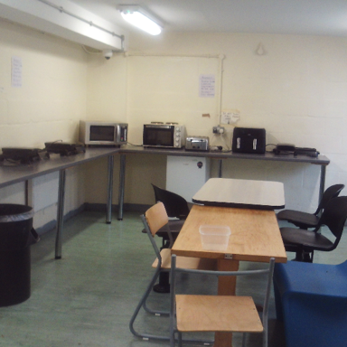 Photograph of the 'reheat room' with microwaves, toasters, air fryers and a trash bin, with beige walls, fluorescent lighting overhead and several tables and chairs arranged in the centre.