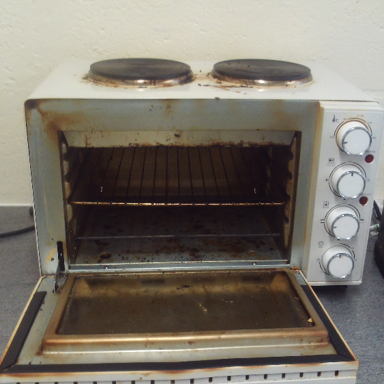 Photograph of a small, white countertop oven with two electric burners on top and an open front door revealing two metal racks inside. Oven shows signs of heavy use with visible rust and grime, and four control knobs on the right side for temperature and function settings.