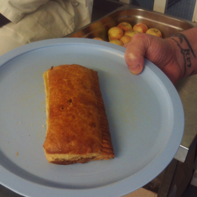 Photograph showing a hand holding a blue plate with a rectangular baked pastry on it. In the background, a tray filled with apples and a person wearing a white coat are visible.