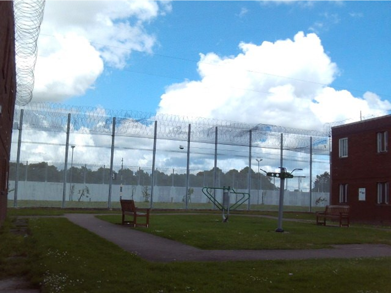 Photograph of an outdoor prison yard enclosed by tall fences topped with barbed wire under a partly cloudy sky. The yard contains benches, a paved walking path, and exercise equipment, with a red brick building visible on the right side.