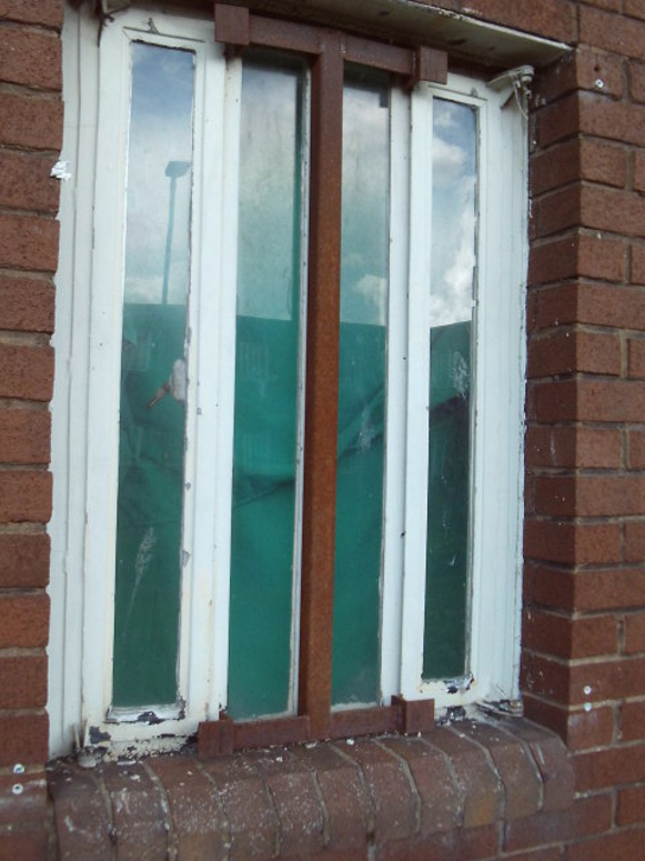 Photograph of a narrow, vertically segmented window set in a red brick wall, featuring four glass panels separated by white and brown frames. The window shows signs of wear with peeling paint and dirt along the bottom edges.