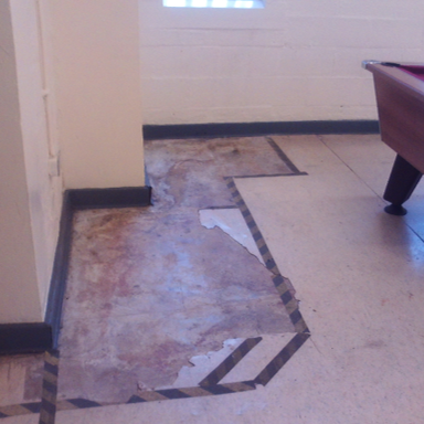 Photograph showing a partially removed floor covering revealing worn and stained subfloor beneath. A pool table with dark legs and red felt is positioned on remaining intact flooring adjacent to the exposed area, which is bordered by black and white striped tape.