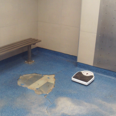 Photograph of a small room with blue flooring, a wooden bench mounted on the wall, and a white scale on the floor. The floor has a large worn patch revealing underlying material and visible cracks, suggesting heavy use or damage.