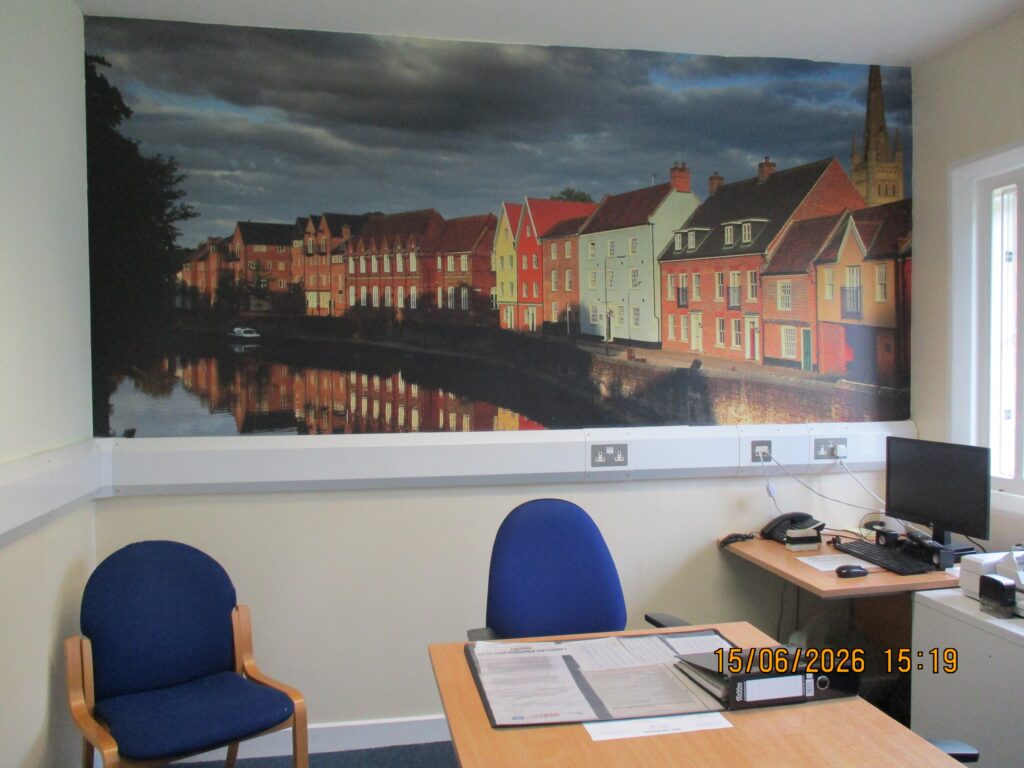Photograph of the adjudication room. It has a table in the middle with two chairs , and to one side is a desk with a computer. On the rear wall there is a large artwork featuring many houses along a river towpath.