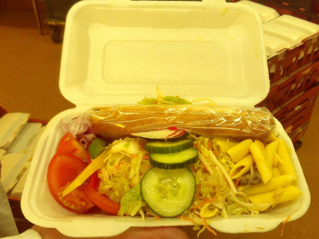 A meal in a takeaway container, featuring various items including cucumber and tomato.