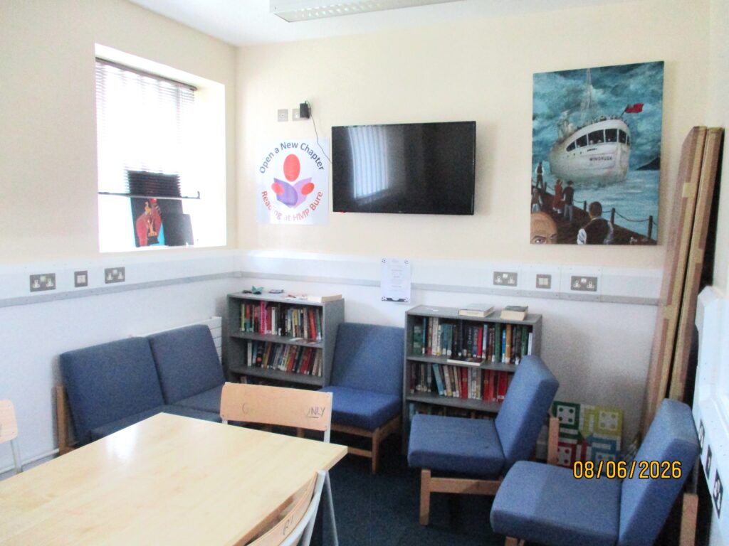 Photograph of the group room for prisoner-led activities. There is a table in the middle with dining chairs around it, and around the edge of the room are some easy chairs and a couple od well-stocked bookcases. There is a screen and an artwork on one wall.