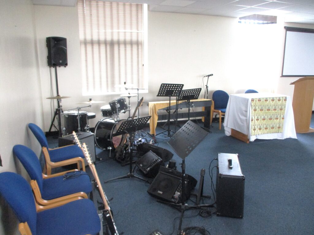 A photograph of the chapel band area. There are some seats around the edge of the room, with music stands, drums, guitars and amplifies arranged next to them.