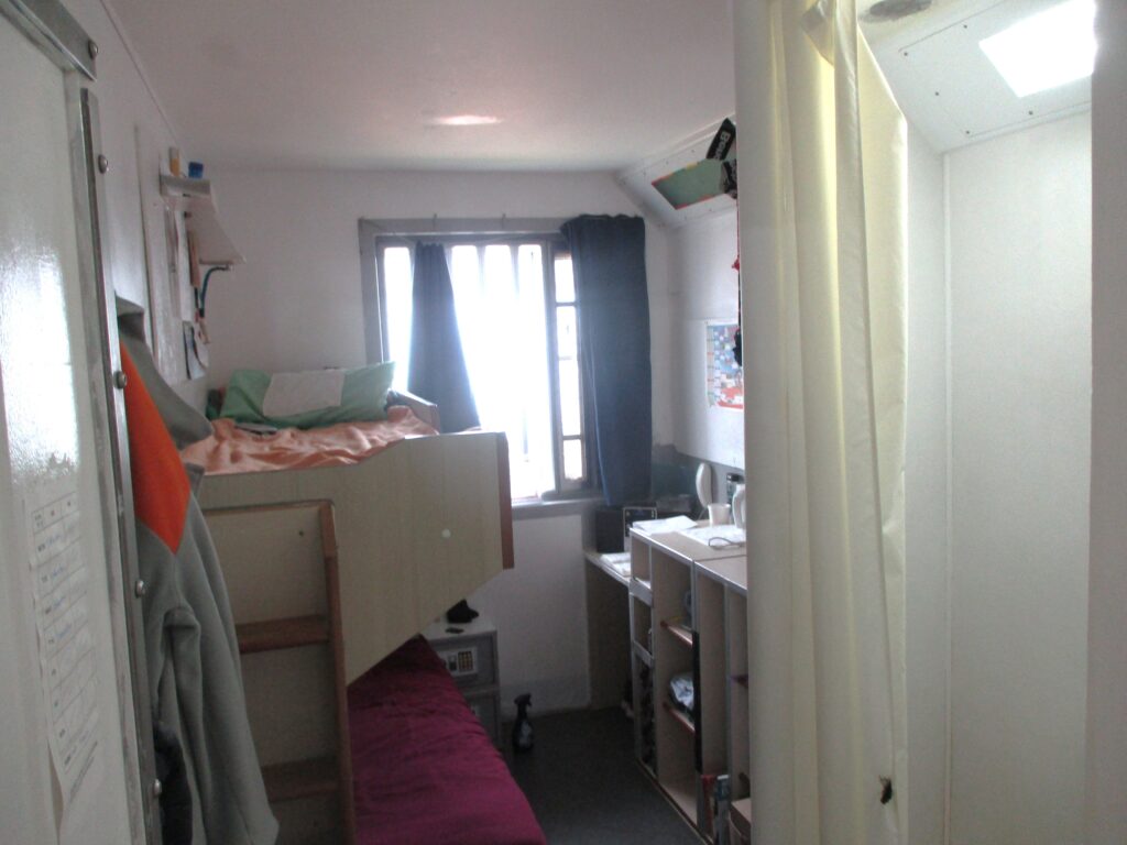 Photograph of a crowded double cell. Bunk beds are one side of the room and other furniture including a desk and are on the other side of the room.