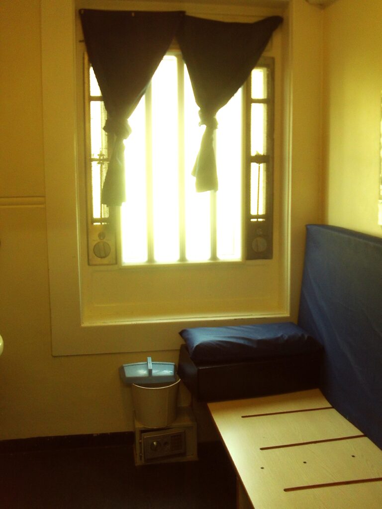 Photograph of a first-night cell, ready for a new occupant. The bed is to one side of the room and sunlight glares in through a window in the back wall.