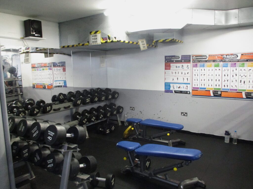 Photograph of the weights room in the gym, in which two benches and numerous sets of free weights can be seen.