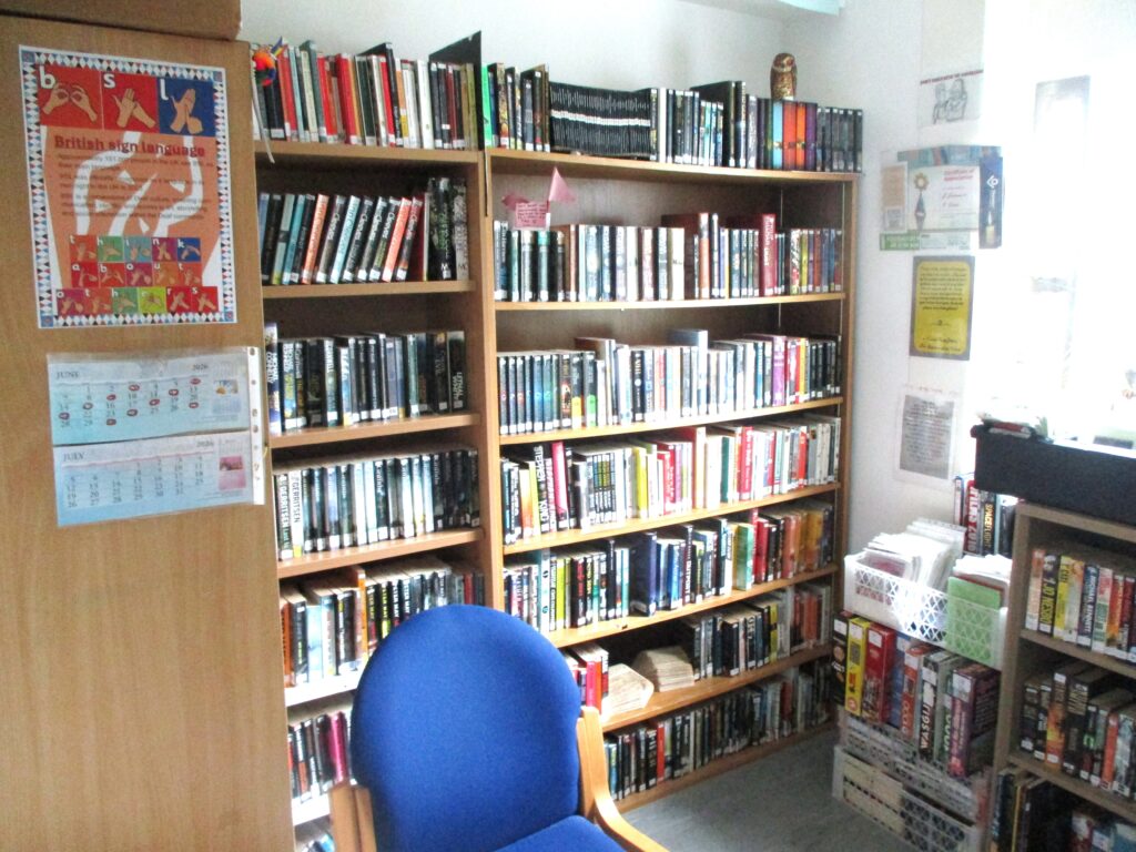 Photograph of an on-wing library on the enabling community unit. Well-stocked bookshelves , psoters with information and a chair can be seen.