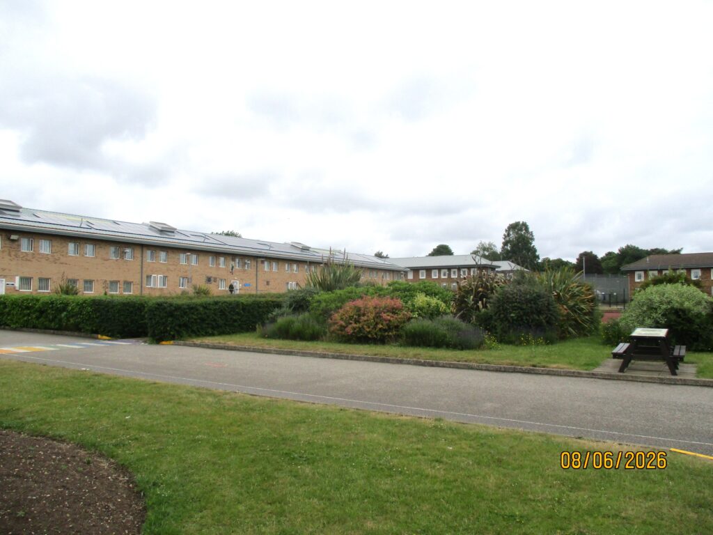 Photograph of an exercise yard, an area with a oath through it and grass and shrubs on either side. Buildings can be seen in the background.