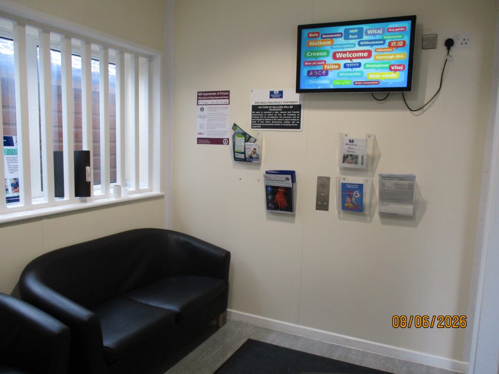 Photograph of the reception holding room. There are some black sofas, racks of information on the wall, and a display screen.