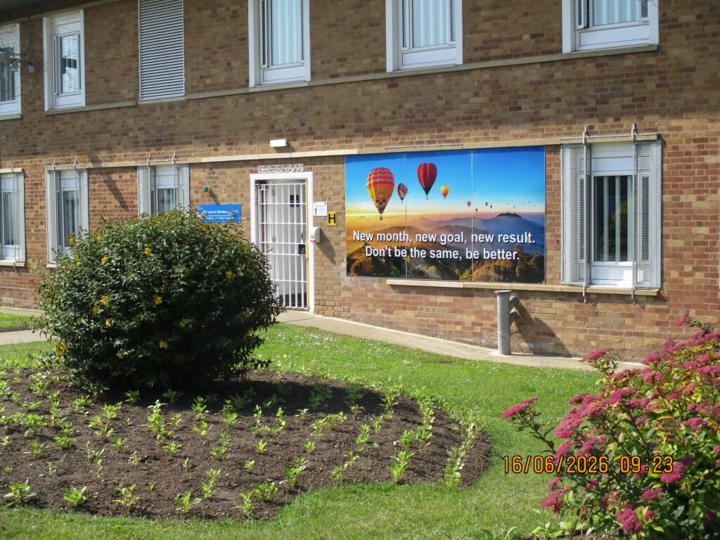 Photograph of the exterior of a residential unit, a brick building with flower beds in front of it. An artwork with a motivational slogan has been installed on the front of the building.